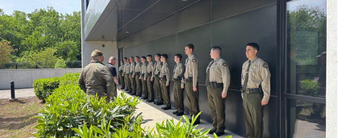 A group of individuals stand at attention outside of a gray, modern building at attention. Two instructors stand in front of them. Green bushes line the foreground.