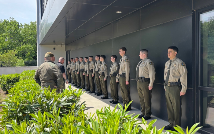 A group of individuals stand at attention outside of a gray, modern building at attention. Two instructors stand in front of them. Green bushes line the foreground.