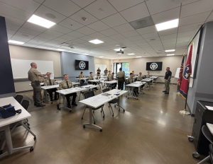 Individuals in uniform sit at attention in a classroom. Two instructors stand in the classroom giving instructions.