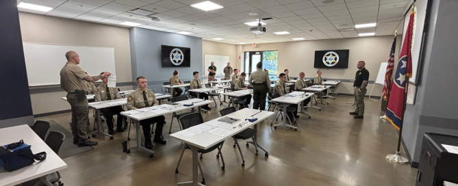 Individuals in uniform sit at attention in a classroom. Two instructors stand in the classroom giving instructions.