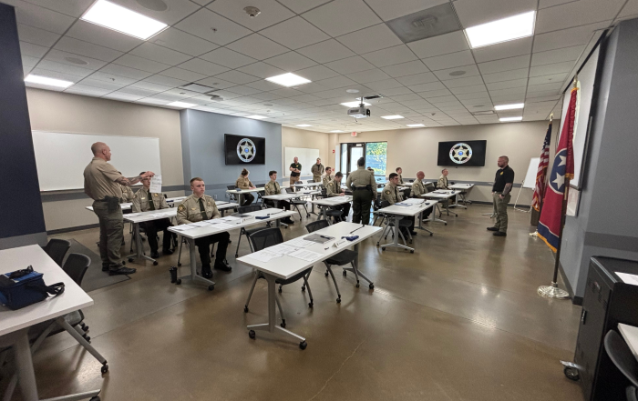 Individuals in uniform sit at attention in a classroom. Two instructors stand in the classroom giving instructions.
