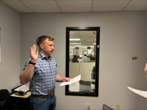 Man with brown hair and mustache, wearing blue jeans and checkered blue shirt, standing in an office with his right hand raised, left hand holding a piece of paper, as he recites an oath.