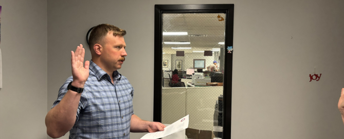 Man with brown hair and mustache, wearing blue jeans and checkered blue shirt, standing in an office with his right hand raised, left hand holding a piece of paper, as he recites an oath.