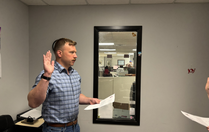 Man with brown hair and mustache, wearing blue jeans and checkered blue shirt, standing in an office with his right hand raised, left hand holding a piece of paper, as he recites an oath.