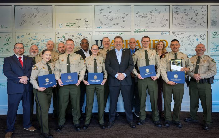 Williamson County Sheriff’s Office deputies, command staff members stand together at the Tennessee Law Enforcement Training Academy graduation. Several deputies in uniform hold blue TLETA certificate folders, with signed graduation boards displayed on the wall behind them.