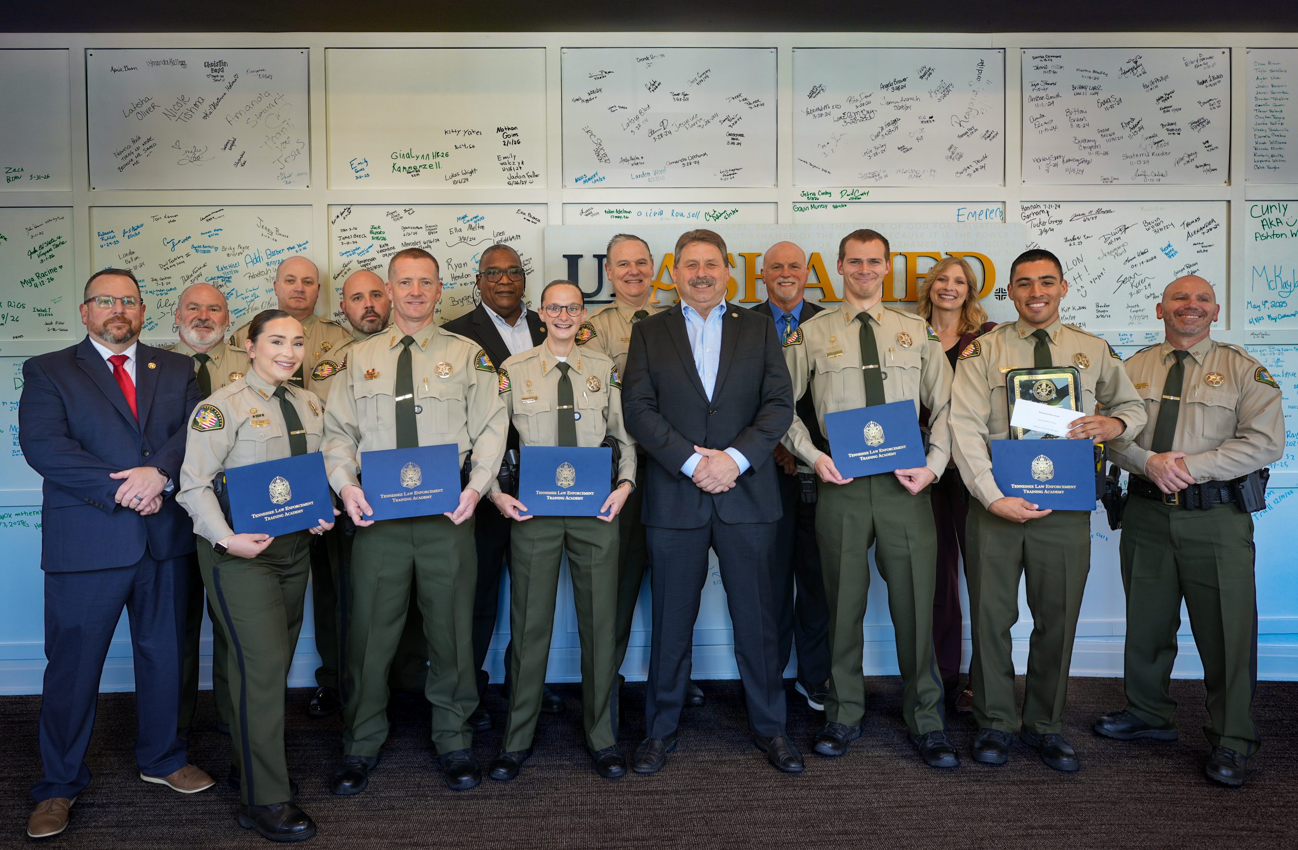 Williamson County Sheriff’s Office deputies, command staff members stand together at the Tennessee Law Enforcement Training Academy graduation. Several deputies in uniform hold blue TLETA certificate folders, with signed graduation boards displayed on the wall behind them.