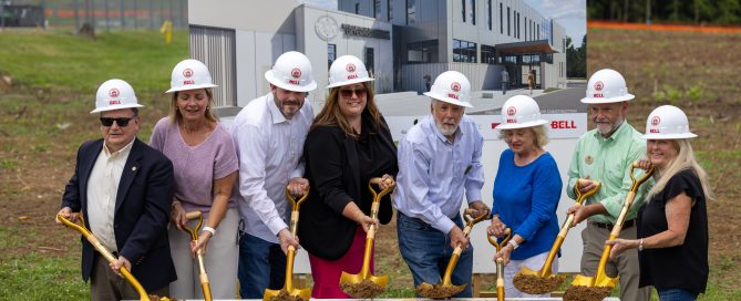 Community leaders, county officials, and project stakeholders gather for the groundbreaking ceremony of the new Williamson County Adult Detention Center. Eight participants wearing white hard hats hold gold ceremonial shovels over a display box filled with soil. A large rendering of the future detention center serves as the backdrop to the ceremony.