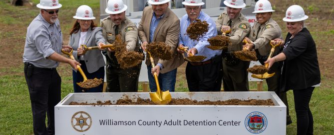 Community leaders, county officials, and project stakeholders gather for the groundbreaking ceremony of the new Williamson County Adult Detention Center. Eight participants wearing white hard hats hold gold ceremonial shovels over a display box filled with soil. A large rendering of the future detention center serves as the backdrop to the ceremony.