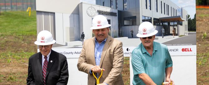 Williamson County officials participate in a ceremonial groundbreaking for the new Williamson County Adult Detention Center. Three men, (former Sheriff, Commisioner Jeff Long, current Sheriff Jeff Hughes, and former Sheriff Ricky Headley) wearing white hard hats stand behind gold ceremonial shovels in a soil-filled display box bearing the Williamson County Sheriff's Office seal, county seal, and project partner logos. Behind them is a large rendering of the future detention center facility.