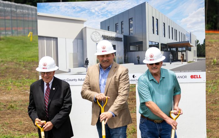 Williamson County officials participate in a ceremonial groundbreaking for the new Williamson County Adult Detention Center. Three men, (former Sheriff, Commisioner Jeff Long, current Sheriff Jeff Hughes, and former Sheriff Ricky Headley) wearing white hard hats stand behind gold ceremonial shovels in a soil-filled display box bearing the Williamson County Sheriff's Office seal, county seal, and project partner logos. Behind them is a large rendering of the future detention center facility.