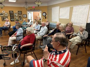 Older adults sit in rows of chairs in a community room, listening to a presentation. Several walkers and mobility aids are visible, and large activity posters hang on the wall behind them.