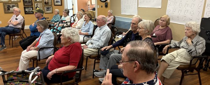 Older adults sit in rows of chairs in a community room, listening to a presentation. Several walkers and mobility aids are visible, and large activity posters hang on the wall behind them.