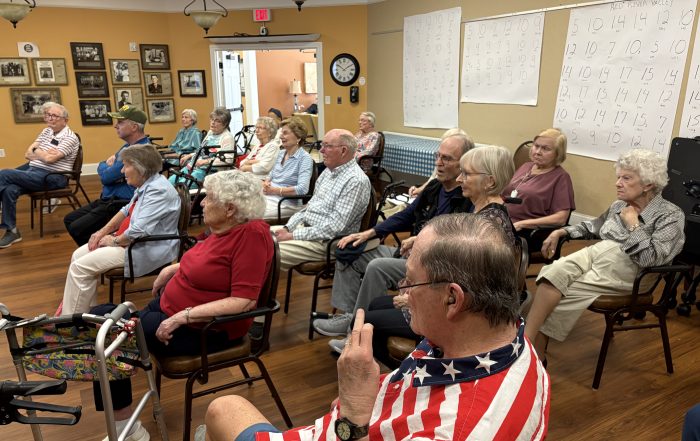 Older adults sit in rows of chairs in a community room, listening to a presentation. Several walkers and mobility aids are visible, and large activity posters hang on the wall behind them.