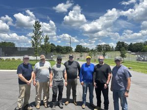 Seven members of the Williamson County Sheriff's Office Reserve Unit stand together on a paved lot outside the Special Operations facility on a sunny day. The volunteers are dressed in work clothes and sunglasses after completing a tree and brush clearing project. Behind them are manicured grounds, security fencing, and a clear blue sky filled with large white clouds. The volunteer effort helped improve safety around the Aviation Unit's helicopter landing area while saving taxpayers thousands of dollars in labor and equipment costs.