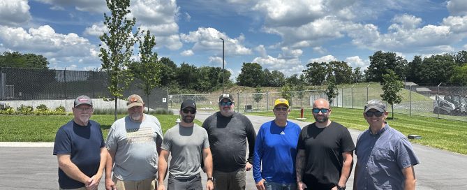 Seven members of the Williamson County Sheriff's Office Reserve Unit stand together on a paved lot outside the Special Operations facility on a sunny day. The volunteers are dressed in work clothes and sunglasses after completing a tree and brush clearing project. Behind them are manicured grounds, security fencing, and a clear blue sky filled with large white clouds. The volunteer effort helped improve safety around the Aviation Unit's helicopter landing area while saving taxpayers thousands of dollars in labor and equipment costs.