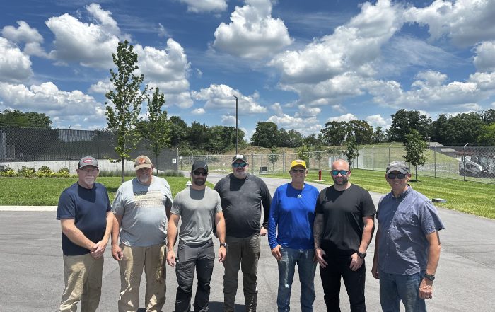 Seven members of the Williamson County Sheriff's Office Reserve Unit stand together on a paved lot outside the Special Operations facility on a sunny day. The volunteers are dressed in work clothes and sunglasses after completing a tree and brush clearing project. Behind them are manicured grounds, security fencing, and a clear blue sky filled with large white clouds. The volunteer effort helped improve safety around the Aviation Unit's helicopter landing area while saving taxpayers thousands of dollars in labor and equipment costs.