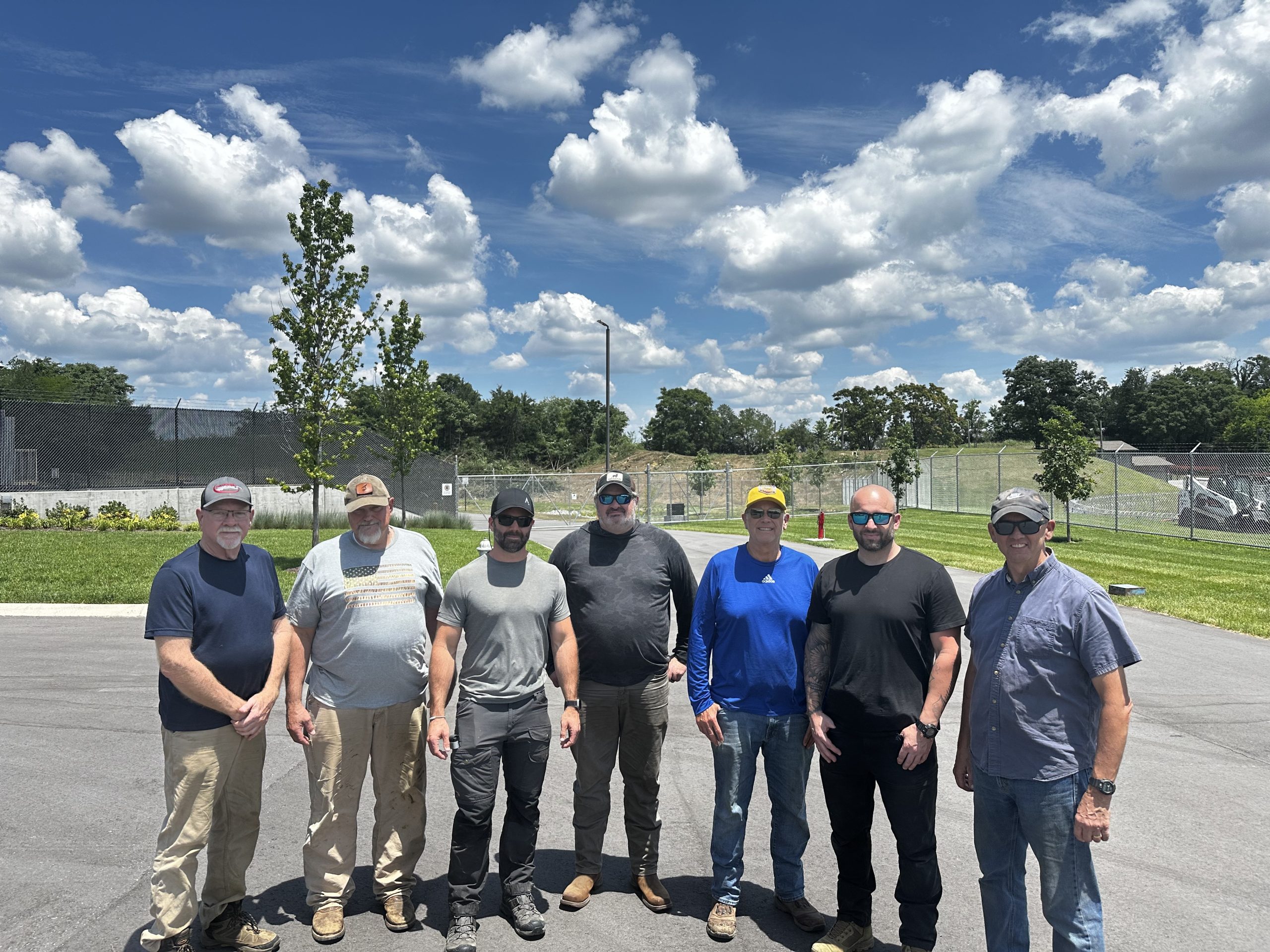 Seven members of the Williamson County Sheriff's Office Reserve Unit stand together on a paved lot outside the Special Operations facility on a sunny day. The volunteers are dressed in work clothes and sunglasses after completing a tree and brush clearing project. Behind them are manicured grounds, security fencing, and a clear blue sky filled with large white clouds. The volunteer effort helped improve safety around the Aviation Unit's helicopter landing area while saving taxpayers thousands of dollars in labor and equipment costs.