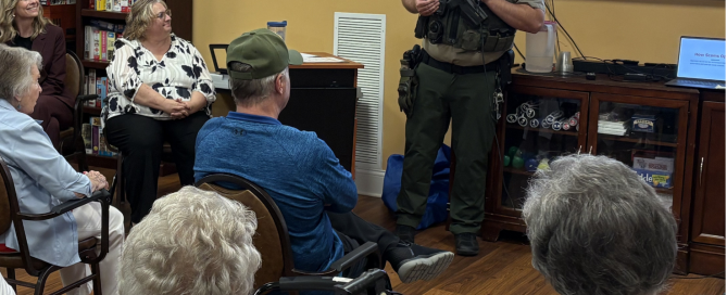 A Williamson County Sheriff’s Office deputy speaks to a group of senior residents during a fraud prevention presentation. A screen behind him displays the slide title, “How Scams Operate,” while attendees listen from their seats.