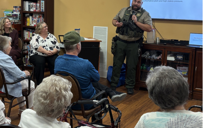 A Williamson County Sheriff’s Office deputy speaks to a group of senior residents during a fraud prevention presentation. A screen behind him displays the slide title, “How Scams Operate,” while attendees listen from their seats.