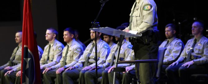 A man in a uniform speaks at a podium while other uniformed individuals sit in chairs behind him, with a Tennessee flag in the foreground.