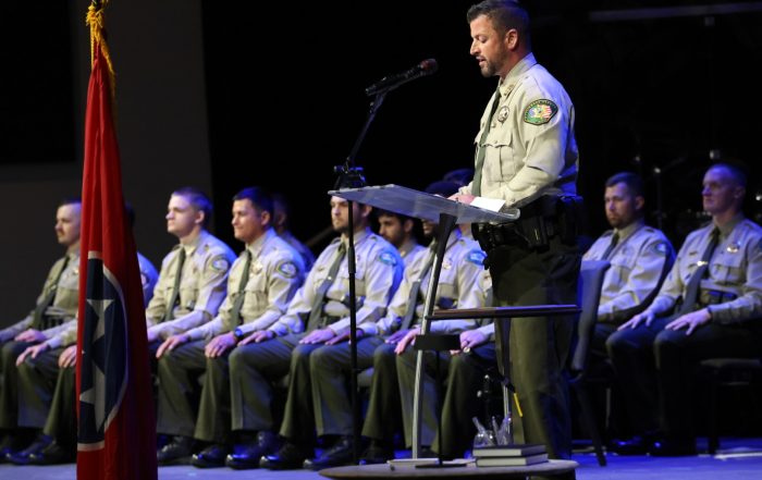 A man in a uniform speaks at a podium while other uniformed individuals sit in chairs behind him, with a Tennessee flag in the foreground.