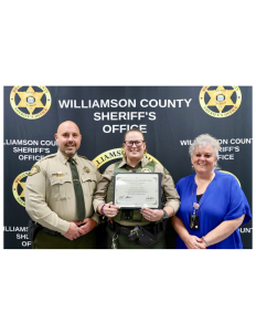 Three people stand in front of a "Williamson County Sheriff's Office" banner. The person in the center, wearing a sheriff's uniform, holds a certificate. The other two individuals, also in uniform and civilian attire, stand on either side of the certificate holder.