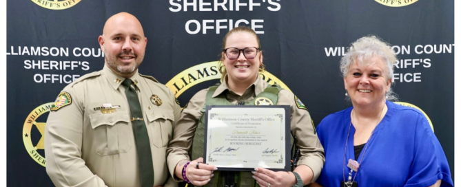 Three people stand in front of a "Williamson County Sheriff's Office" banner. The person in the center, wearing a sheriff's uniform, holds a certificate. The other two individuals, also in uniform and civilian attire, stand on either side of the certificate holder.