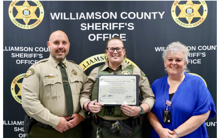 Three people stand in front of a "Williamson County Sheriff's Office" banner. The person in the center, wearing a sheriff's uniform, holds a certificate. The other two individuals, also in uniform and civilian attire, stand on either side of the certificate holder.