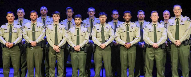 A group of individuals in uniform stand in two rows on a stage with a dark backdrop.