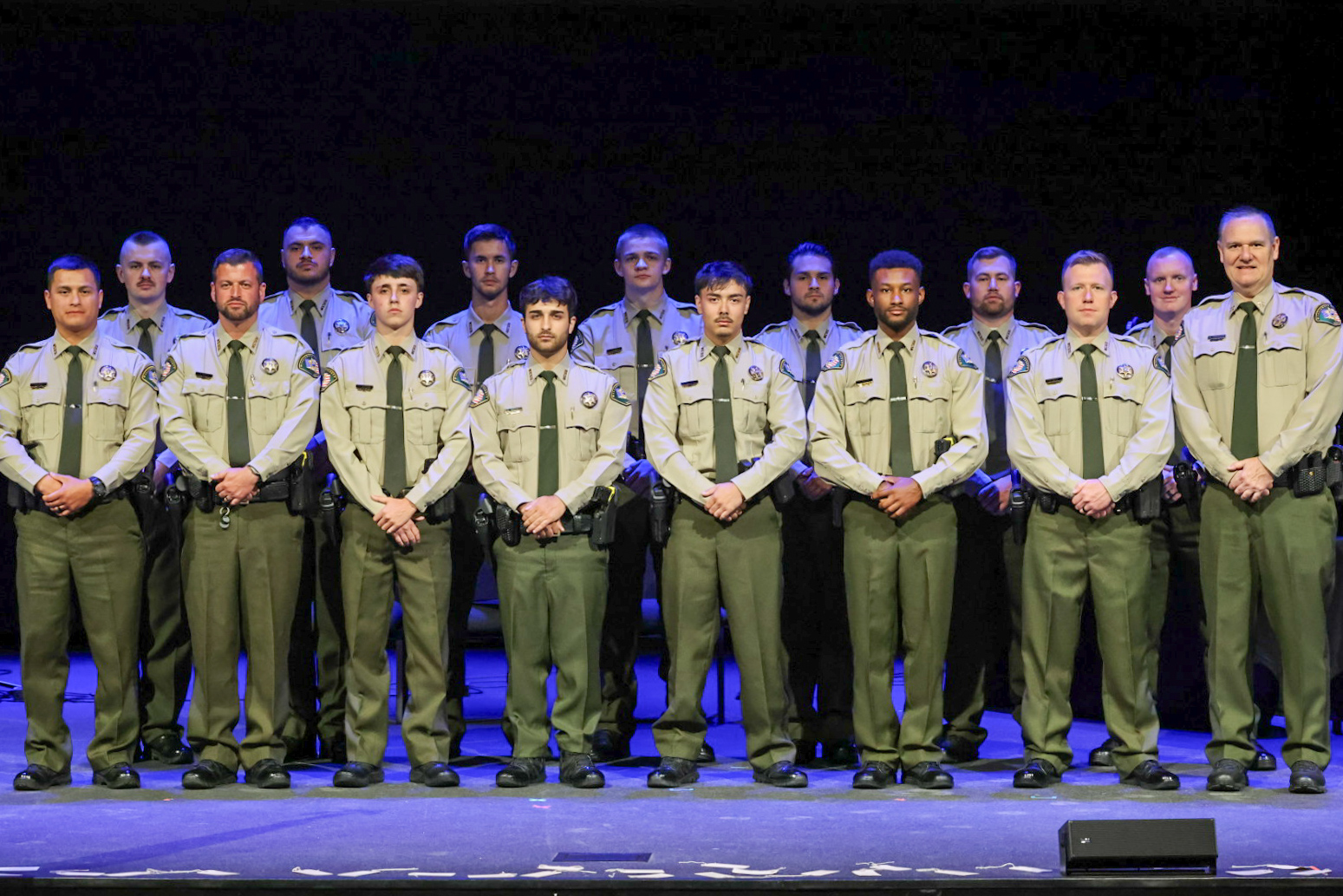 A group of individuals in uniform stand in two rows on a stage with a dark backdrop.