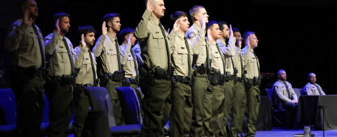 A group of uniformed individuals stand with their right hands raised in an oath during a ceremony. They are wearing light-colored shirts and dark pants, with badges and utility belts. A sheriff's office logo is visible on a screen in the background.
