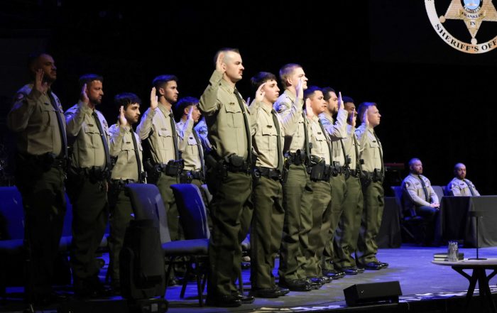 A group of uniformed individuals stand with their right hands raised in an oath during a ceremony. They are wearing light-colored shirts and dark pants, with badges and utility belts. A sheriff's office logo is visible on a screen in the background.