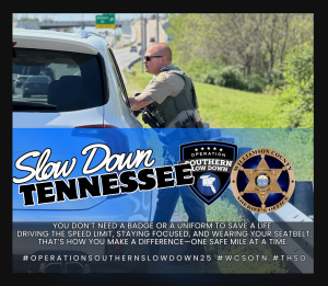 A Williamson County Sheriff's Office deputy speaks with a driver during a traffic stop beside a highway. The graphic reads "Slow Down Tennessee" and features the Operation Slow Down and WCSO Logos. Additional text encourages drivers to obey the speed limit, stay focused, and wear a seatbelt to help save lives.