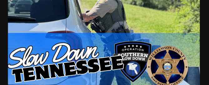 A Williamson County Sheriff's Office deputy speaks with a driver during a traffic stop beside a highway. The graphic reads "Slow Down Tennessee" and features the Operation Slow Down and WCSO Logos. Additional text encourages drivers to obey the speed limit, stay focused, and wear a seatbelt to help save lives.