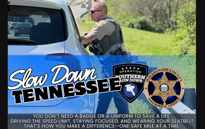A Williamson County Sheriff's Office deputy speaks with a driver during a traffic stop beside a highway. The graphic reads "Slow Down Tennessee" and features the Operation Slow Down and WCSO Logos. Additional text encourages drivers to obey the speed limit, stay focused, and wear a seatbelt to help save lives.