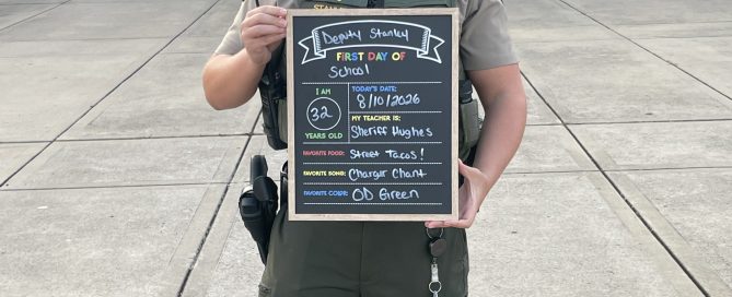 WCSO Deputy Stanley stands outside a school in uniform and sunglasses, holding a “First Day of School” chalkboard sign listing his age as 32, Sheriff Hughes as his teacher, street tacos as his favorite food, Charger Chant as his favorite song, and OD Green as his favorite color.