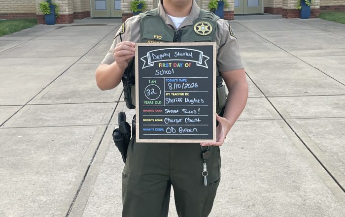 WCSO Deputy Stanley stands outside a school in uniform and sunglasses, holding a “First Day of School” chalkboard sign listing his age as 32, Sheriff Hughes as his teacher, street tacos as his favorite food, Charger Chant as his favorite song, and OD Green as his favorite color.