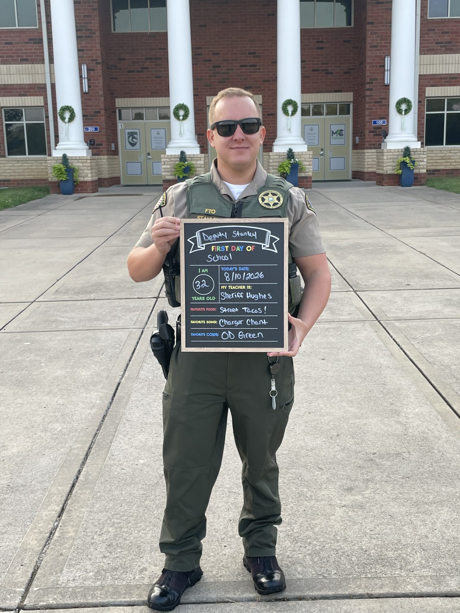 WCSO Deputy Stanley stands outside a school in uniform and sunglasses, holding a “First Day of School” chalkboard sign listing his age as 32, Sheriff Hughes as his teacher, street tacos as his favorite food, Charger Chant as his favorite song, and OD Green as his favorite color.