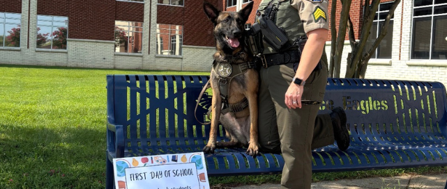 Sgt. Amy Fann stands beside WCSO K9 Nala outside Independence High School on the first day of school. Nala is sitting on a blue Eagles bench beneath blooming trees, with a colorful first-day-of-school sign displayed in front of them.