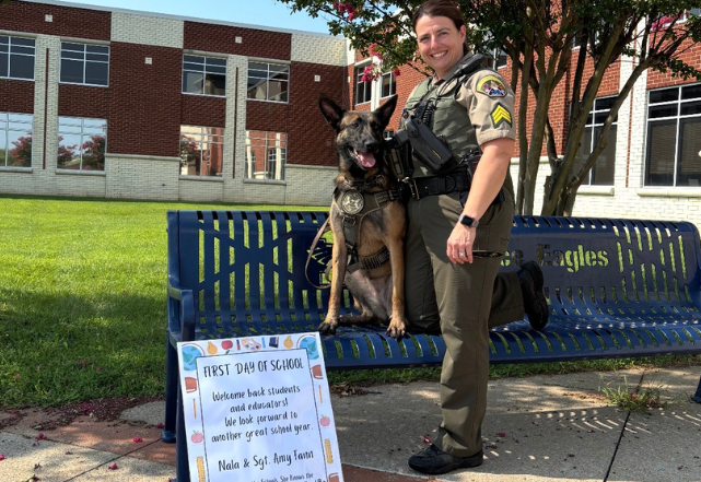 Sgt. Amy Fann stands beside WCSO K9 Nala outside Independence High School on the first day of school. Nala is sitting on a blue Eagles bench beneath blooming trees, with a colorful first-day-of-school sign displayed in front of them.