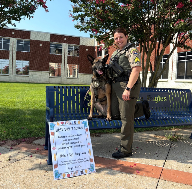 Sgt. Amy Fann stands beside WCSO K9 Nala outside Independence High School on the first day of school. Nala is sitting on a blue Eagles bench beneath blooming trees, with a colorful first-day-of-school sign displayed in front of them.