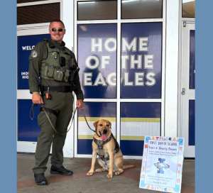 Deputy John Bevans stands with WCSO K9 Scout outside Independence High School on the first day of school. Scout, wearing his K9 harness and badge, sits beside Deputy Bevans in front of the school entrance and a “Home of the Eagles” sign. A colorful first-day-of-school sign is displayed beside Scout.