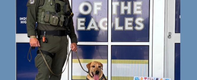 Deputy John Bevans stands with WCSO K9 Scout outside Independence High School on the first day of school. Scout, wearing his K9 harness and badge, sits beside Deputy Bevans in front of the school entrance and a “Home of the Eagles” sign. A colorful first-day-of-school sign is displayed beside Scout.