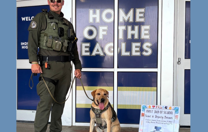 Deputy John Bevans stands with WCSO K9 Scout outside Independence High School on the first day of school. Scout, wearing his K9 harness and badge, sits beside Deputy Bevans in front of the school entrance and a “Home of the Eagles” sign. A colorful first-day-of-school sign is displayed beside Scout.