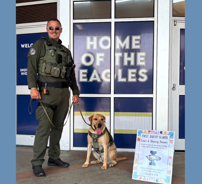 Deputy John Bevans stands with WCSO K9 Scout outside Independence High School on the first day of school. Scout, wearing his K9 harness and badge, sits beside Deputy Bevans in front of the school entrance and a “Home of the Eagles” sign. A colorful first-day-of-school sign is displayed beside Scout.