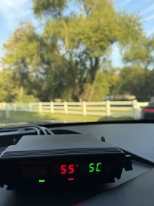 View from inside a patrol vehicle showing a Stalker Dual DSR radar unit on the dashboard. The radar display shows a target speed of 55 mph and a patrol speed of 52 mph, with a tree-lined roadway and white fence visible through the windshield.