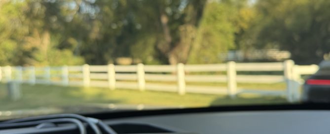 View from inside a patrol vehicle showing a Stalker Dual DSR radar unit on the dashboard. The radar display shows a target speed of 55 mph and a patrol speed of 52 mph, with a tree-lined roadway and white fence visible through the windshield.