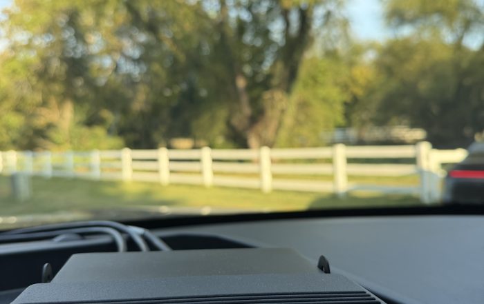 View from inside a patrol vehicle showing a Stalker Dual DSR radar unit on the dashboard. The radar display shows a target speed of 55 mph and a patrol speed of 52 mph, with a tree-lined roadway and white fence visible through the windshield.