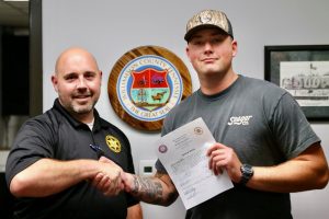 Capt. Chad Youker, left, shakes hands with newly sworn-in Detention Deputy Blake Larson, right, who holds his oath of office. The Williamson County, Tennessee, seal is displayed on the wall behind them.