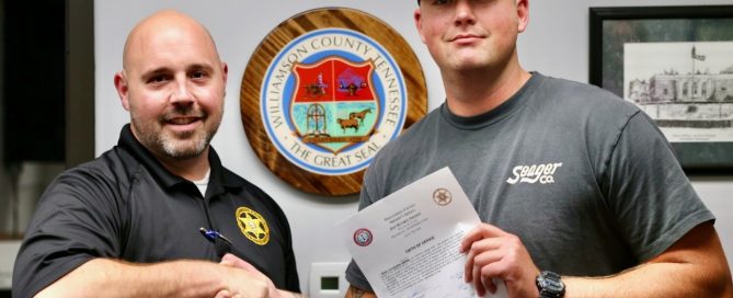Capt. Chad Youker, left, shakes hands with newly sworn-in Detention Deputy Blake Larson, right, who holds his oath of office. The Williamson County, Tennessee, seal is displayed on the wall behind them.